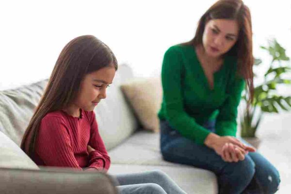 A child sits looking down while an adult leans toward them from a nearby sofa